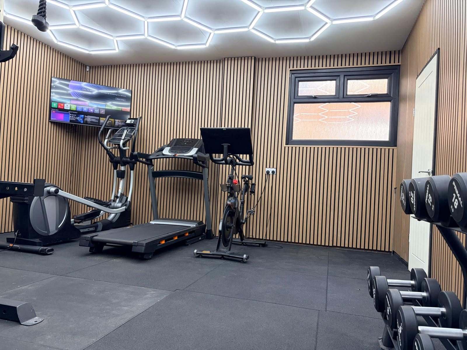 Cardio end of the Durham garage gym with cross trainer, treadmill and exercise bike under hexagon LED lights, with a wall-mounted TV and new window
