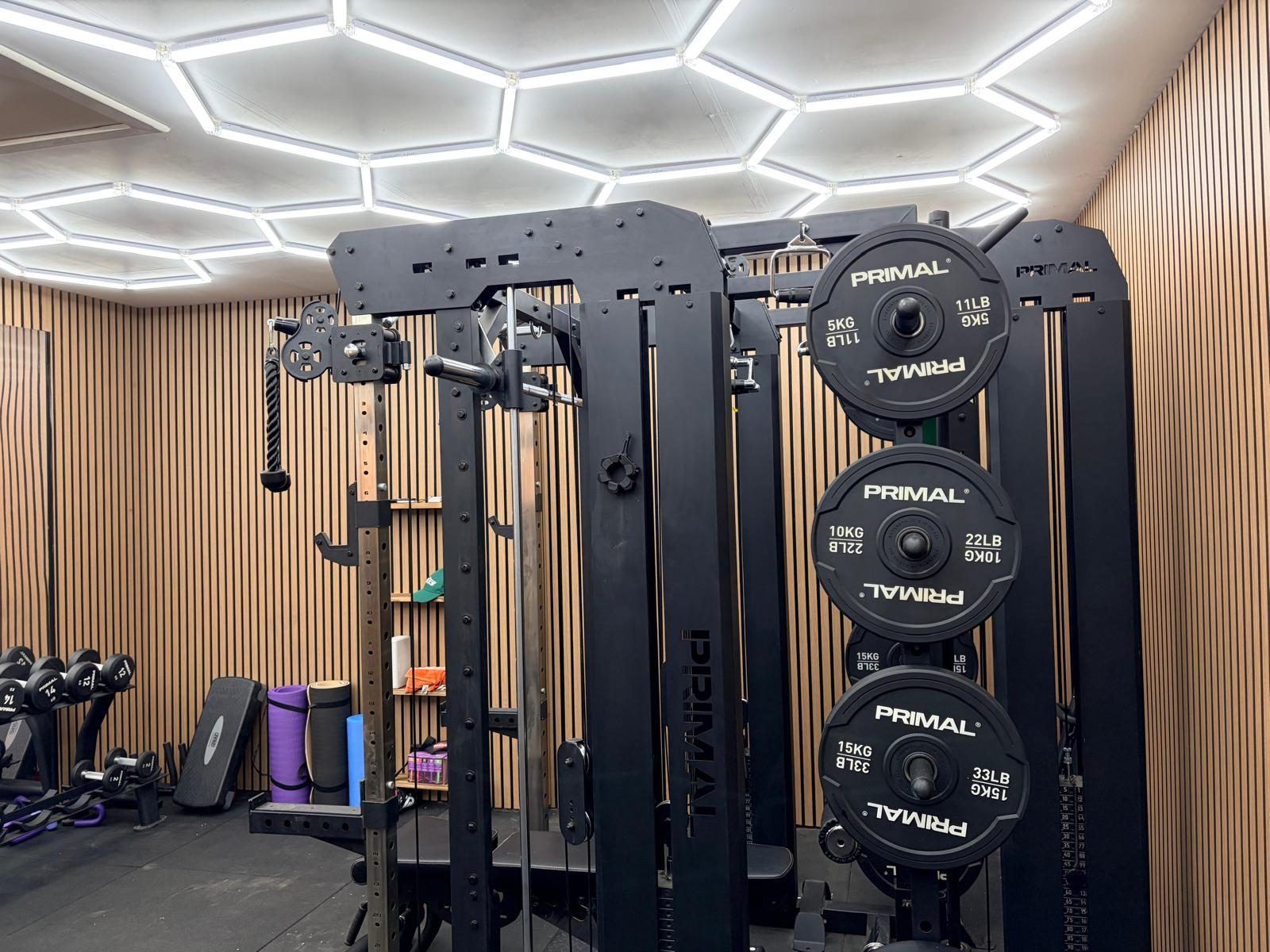 Close view of a black multi-station cage rack loaded with weight plates, with mats and storage shelving behind on wood slat panelling