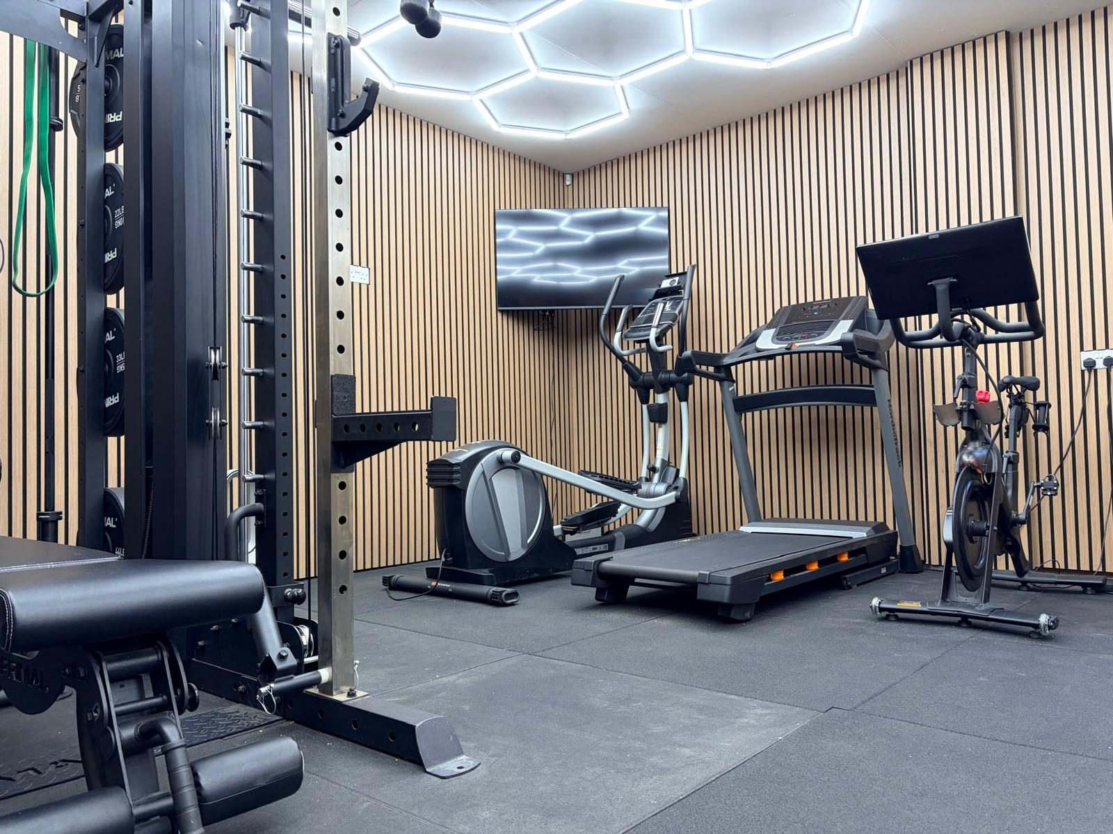 View past the cable column of the cage rack towards the cardio machines and TV under the hexagon LED ceiling