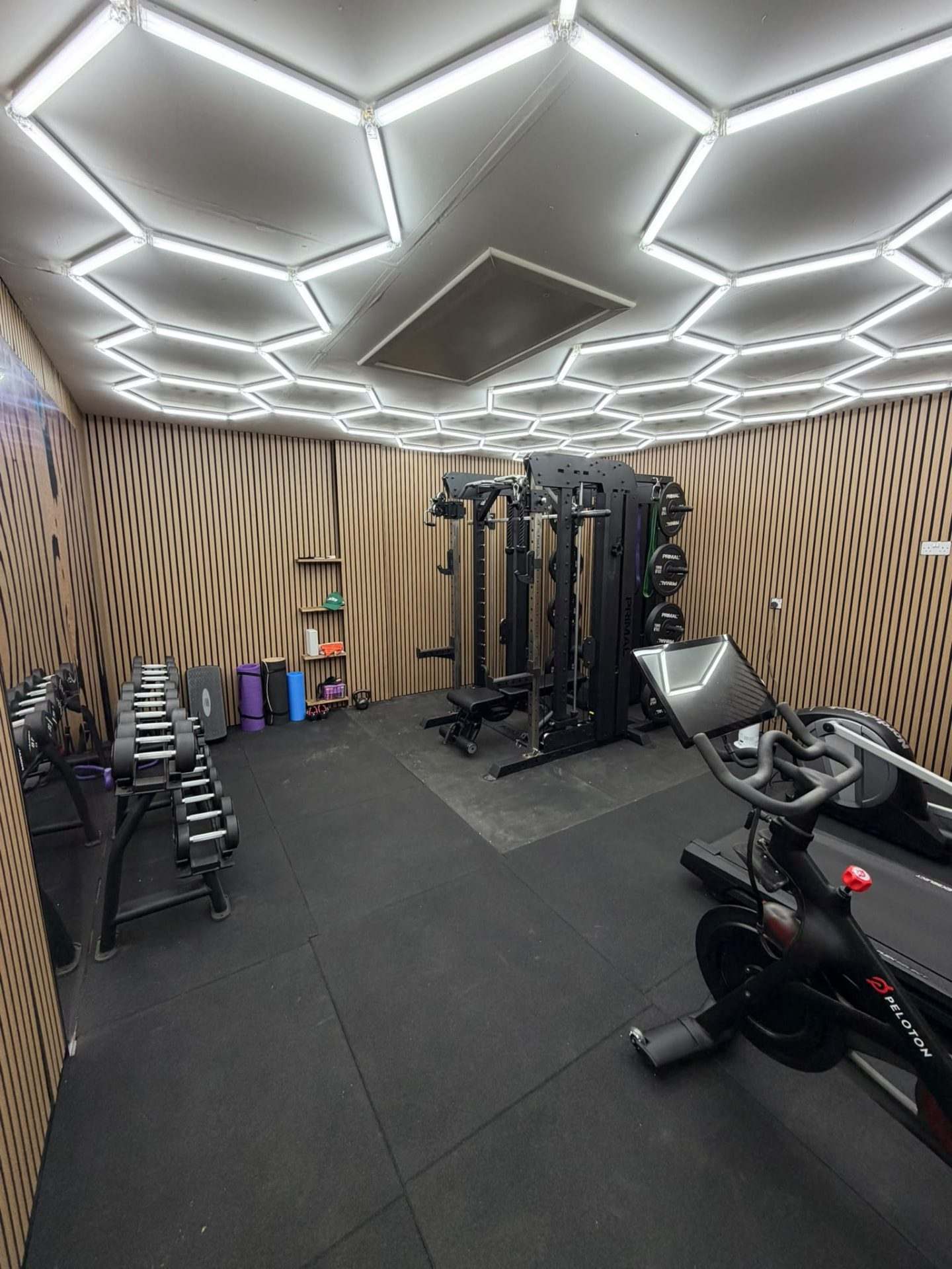 Wide view down the double garage gym showing the hexagon LED ceiling, dumbbell rack, cage rack and exercise bike